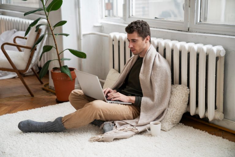 أنظمة التدفئة المركزية في مصر - Central Heating system in egypt Man working on a laptop near a radiator, wrapped in a blanket, illustrating a cozy indoor environment with heating systems.